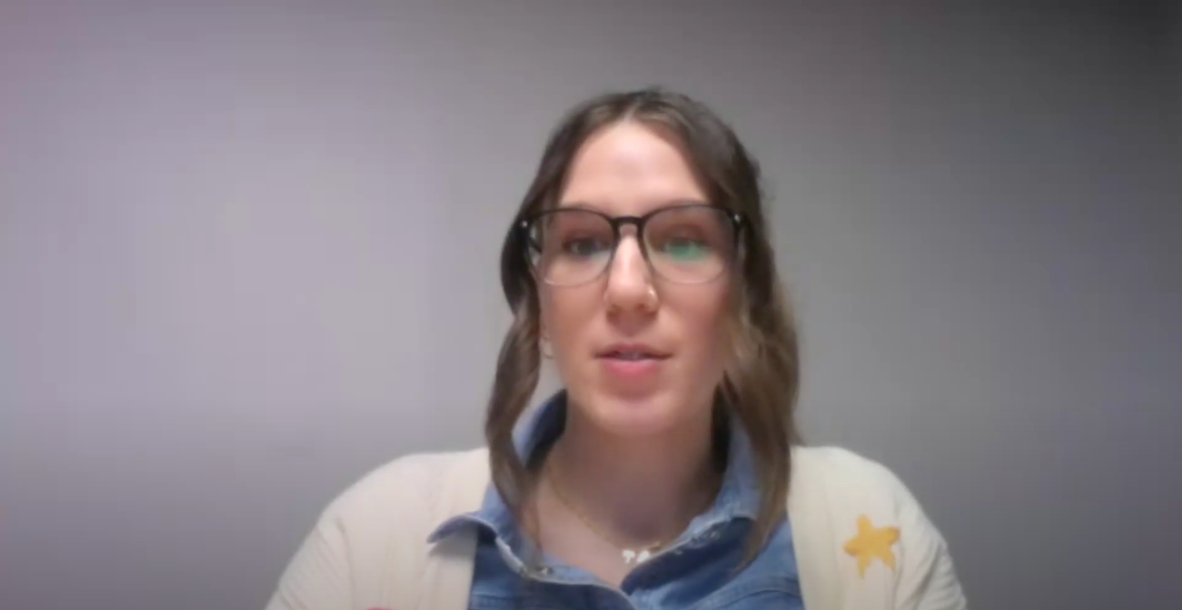 A woman with light brown hair in loose waves, wearing black-rimmed glasses, a denim shirt, and a cream-colored cardigan with a yellow star patch, speaks during a video call. She is in front of a plain gray wall, and her facial expression is engaged and focused. The lighting is soft and even.